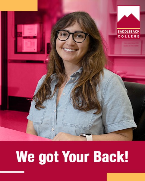Female student with glasses standing in a hallway smiling at the camera. We got your back!