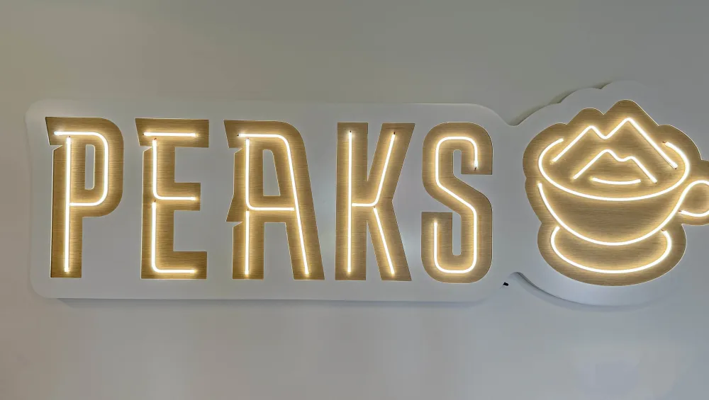Neon sign that reads PEAKS with a mug near it filed with coffee and where the foam is shaped into peaks.