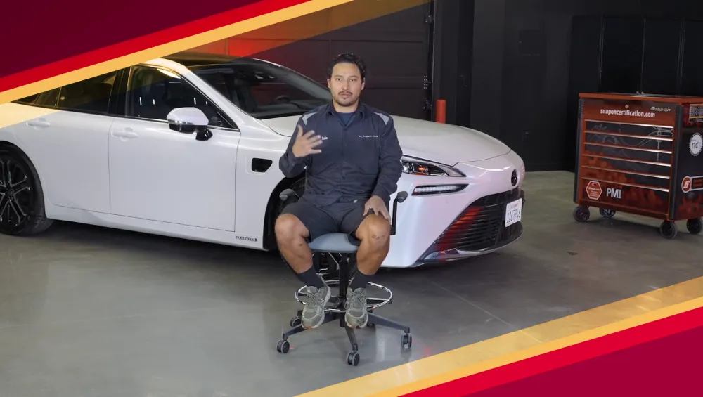 Male student sitting being interviewed in front of a white car in the autobay.