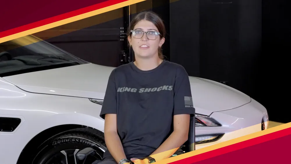 Female student sitting in front of white car being interviewed.