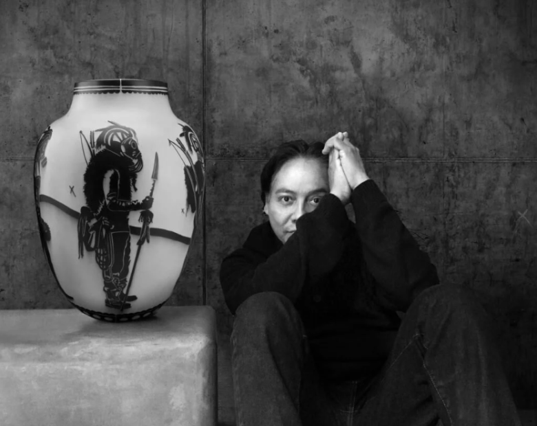 Black and white image. Artist Virgil Ortiz sits, in a black shirt and jeans with his hands together, next one of his pieces, which is an oval-shaped piece of pottery featuring a Native American warrior.