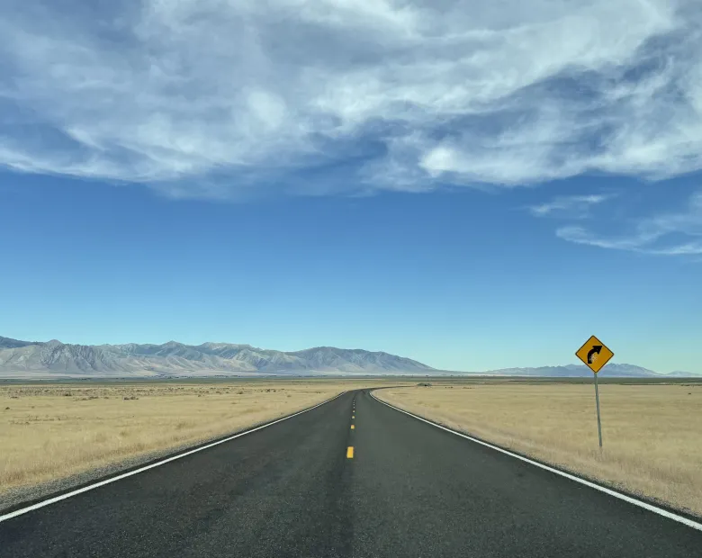 A two lane highway stretching into the distance in the middle of a barren desert with mountains in the background and a right turn yellow traffic sign at the side of the road.