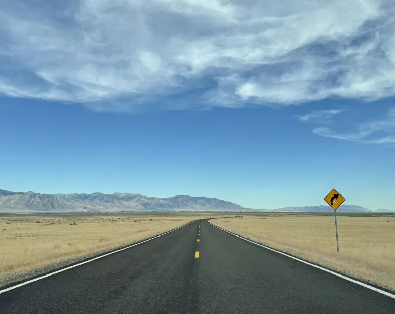A two lane highway stretching into the distance in the middle of a barren desert with mountains in the background and a right turn yellow traffic sign at the side of the road.