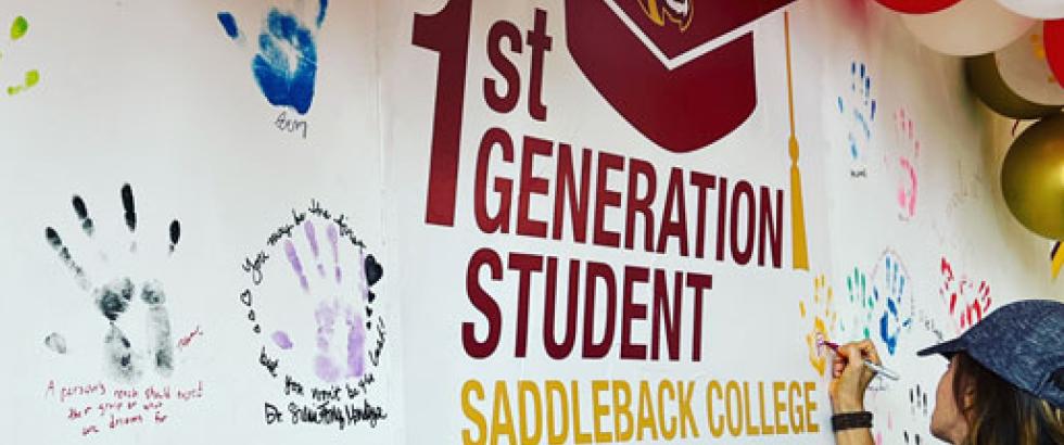 A wall with the words 1st Generation Student Saddleback College is decorated with the painted handprints of students. A female student is pictured writing her name on her handprint.