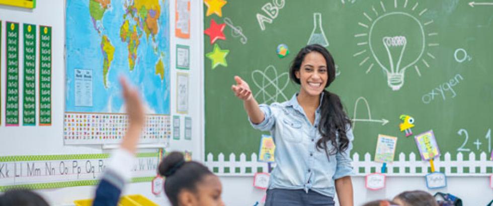 Teacher at the front of the classroom pointing to a student who has their hand raised.