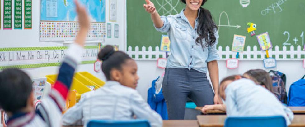 Teacher at the front of the classroom pointing to a student who has their hand raised.
