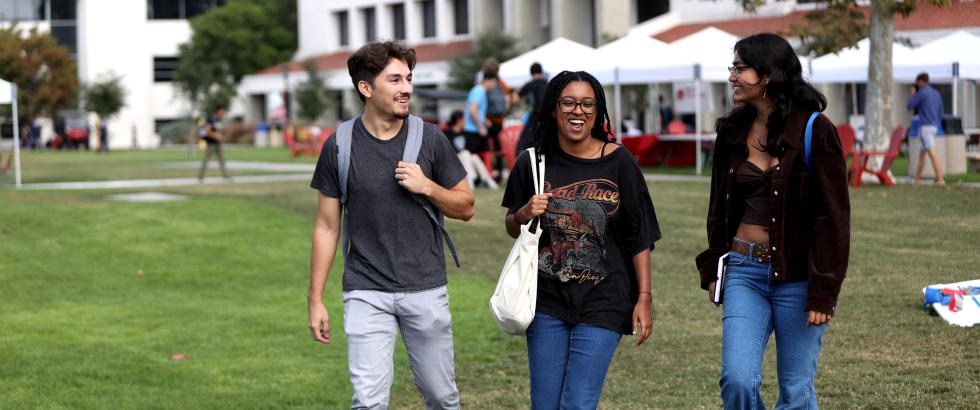 three students walking across a grassy quad with others in the background