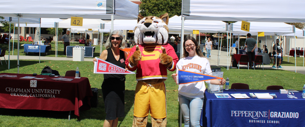 saddleback college mascot, blaze the bobcat, standing in front of two outdoor booths with representatives from colleges