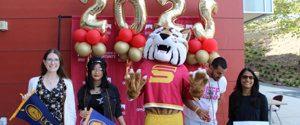 Four students pose with the Saddleback College mascot in front of a red backdrop and gold “2023” balloons at a transfer celebration. Two students hold UC Davis pennants, and one holds a UC Davis sign. Red and gold balloons decorate the display, celebrating the graduates’ transfer achievements.