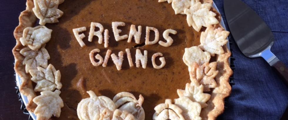 A pumpkin pie decorated with pastry cutouts spelling “Friendsgiving” in the center and surrounded by leaf- and pumpkin-shaped crust pieces along the edge. The pie sits on a blue cloth with a metal pie server beside it.