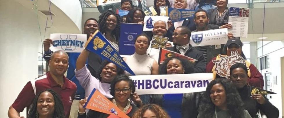 A large, joyful group of people of various ages stands on indoor stairs holding signs and banners representing Historically Black Colleges and Universities (HBCUs). The group displays school pennants for institutions such as Florida A&M, Dillard, Fort Valley State, Cheyney, and Bennett College, along with large signs reading “#HBCUcaravan.” Colorful balloons in green and purple are tied above the group, and the photo is taken in a bright, modern building.