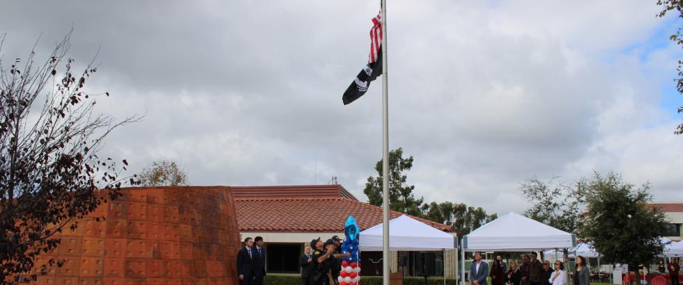 A Veterans Day ceremony at Saddleback College shows uniformed officers saluting as the American and POW/MIA flags are raised on a flagpole. People stand under white canopies watching the event, and red, white, and blue balloon decorations are nearby. The ceremony takes place in front of a curved copper memorial wall under a cloudy sky.