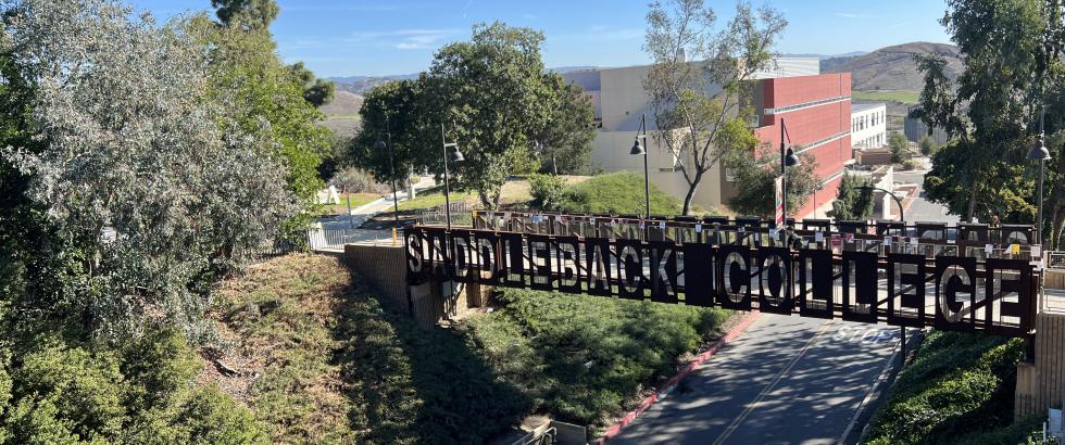 A pedestrian bridge with large metal letters spelling “Saddleback College” spans a campus road. Trees and greenery surround the bridge, and in the background, a red and white campus building sits against rolling hills under a clear blue sky.