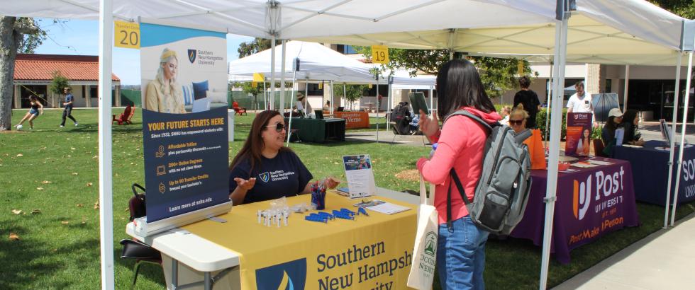 representative wearing sunglasses and an SNHU shirt speaks with a student holding a tote bag and wearing a backpack. The table is covered with promotional items and a yellow SNHU tablecloth. A large display sign behind the representative reads “YOUR FUTURE STARTS HERE” and lists information about affordable tuition, 200+ online degree programs, and transfer credits.