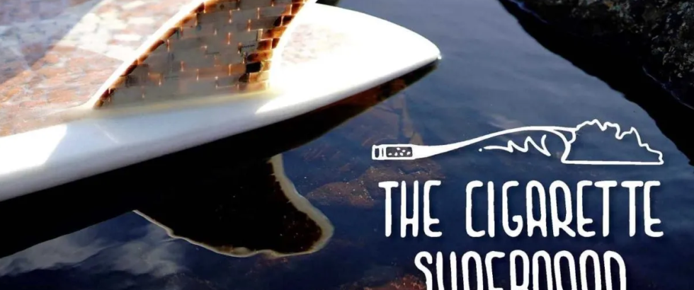 A close-up of a surfboard resting in shallow water beside dark rocks. The board’s fin is made from stacked cigarette butts encased in clear material. The surface of the water reflects the board and sky. Over the water is white stylized text with a small illustration of a cigarette forming a wave line. The text reads: “THE CIGARETTE SURFBOARD” and below it: “A FILM BY BEN JUDKINS.”