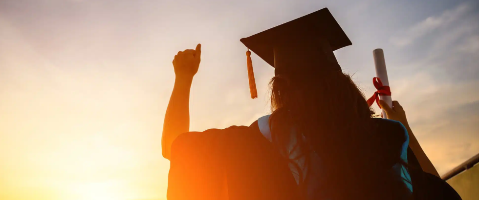 A graduate in a cap and gown stands outdoors, shown from behind, raising one arm in a celebratory gesture while holding a rolled diploma tied with a ribbon in the other hand. The person is silhouetted against a bright, low sun, with soft light outlining their figure.