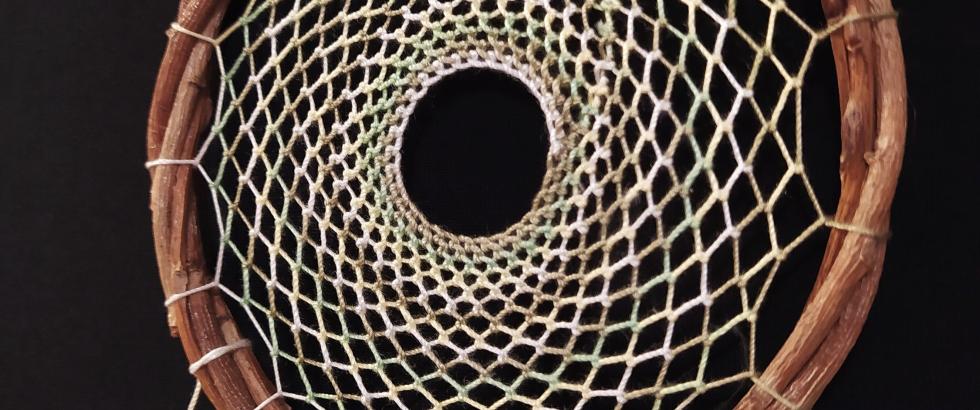 A handcrafted dreamcatcher made with a circular wooden hoop wrapped in twine, featuring an intricate woven web pattern with a hole in the center. Several soft brown and white feathers hang from the bottom, each tied with a small wooden bead. The background is black, highlighting the details.