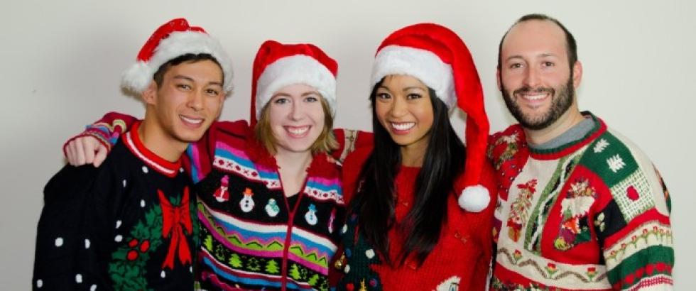 Four adults stand close together, smiling at the camera and wearing festive holiday sweaters and Santa hats. Their sweaters include patterns such as bows, trees, snowmen, animals, and the word “NO.” They pose against a plain background.