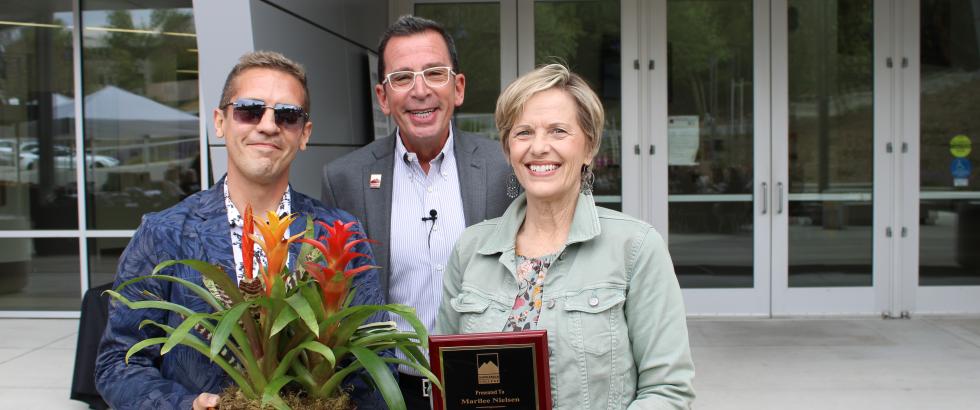 Three adults stand smiling outside a building entrance. The person on the left holds a potted plant with long leaves and tall flower spikes. The person in the center stands behind them, wearing glasses and a microphone. The person on the right holds a plaque that reads: “Presented To Marlene Nielsen. Outstanding Emeritus Professor of the Year. In Recognition Of Exceptional And Influential Teaching, Commitment, And Dedication. 2004–2023.”