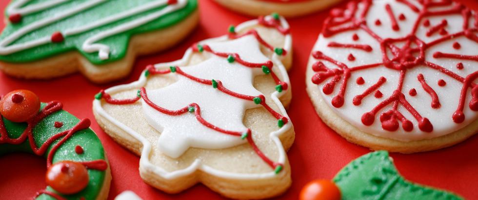 A close-up view of decorated holiday sugar cookies on a red surface, including tree-shaped cookies with white or green icing and garland designs, round cookies with intricate snowflake patterns, a wreath-shaped cookie with small round decorations, and a holly-leaf-shaped cookie with a single round berry.