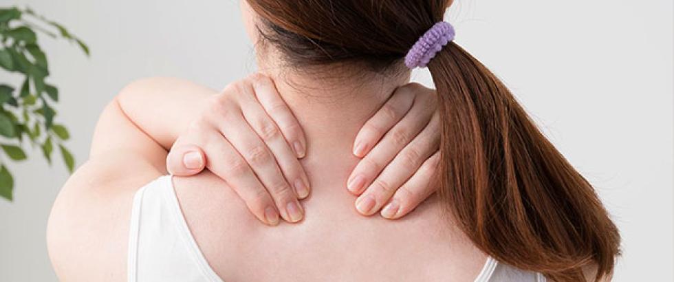 A person with long hair tied back holds both hands on the back of their neck as if massaging or relieving discomfort. The individual is shown from behind, wearing a sleeveless top, with part of a leafy plant visible in the background.