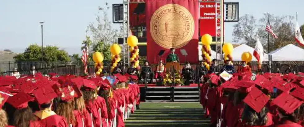 Graduates in caps and gowns sit in rows facing a stage at an outdoor ceremony. A large banner reads “Saddleback College” and “Commencement,” with a seal displayed at center. Balloons flank the stage, and speakers and faculty sit behind a podium