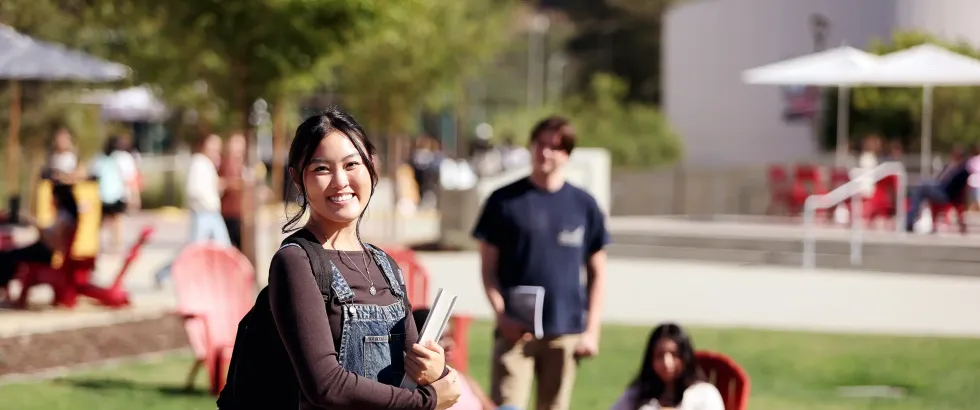 Female Saddleback College student standing with backpack and book with on the quad with students sitting in group in the background.