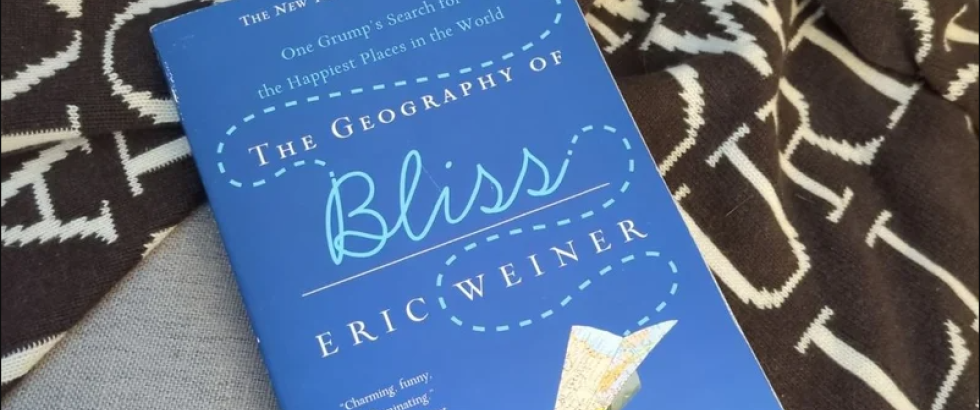 A blue paperback book titled “The Geography of Bliss” by Eric Weiner rests on a gray surface beside a black-and-cream patterned blanket. The cover reads: “The New York Times Bestseller,” “One Grump’s Search for the Happiest Places in the World,” “The Geography of Bliss,” “Eric Weiner,” “Charming, funny, and illuminating. – Washington Post Book World,” and “Moving, profound… a winning book. – National Geographic.” The cover features a hand holding a paper airplane made from a map.