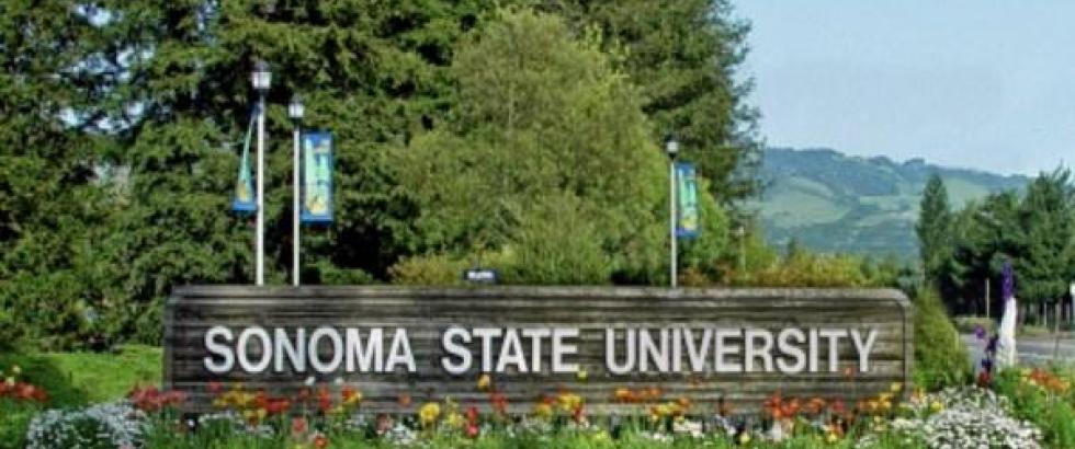 Wooden campus sign reading “Sonoma State University” sits in front of a landscaped bed of colorful flowers. Tall evergreen trees rise behind the sign, with green hills visible in the distance under a clear sky.