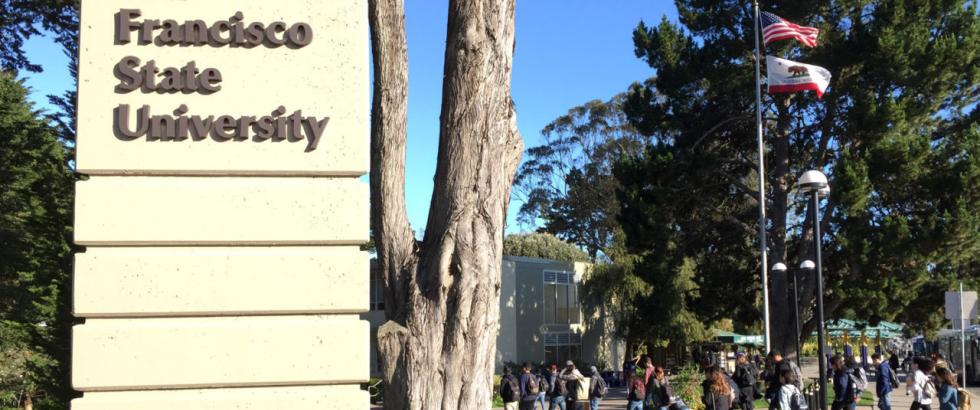 Large campus sign reading “San Francisco State University” stands beside tall trees. In the background, students walk along a wide pathway, and the United States and California state flags fly on flagpoles against a clear blue sky.