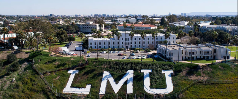 Aerial view of a hillside with large white letters spelling “LMU” in the foreground. Behind the hillside sits a university campus with multi-story academic buildings, palm trees, parked cars, and a city skyline in the distance under a clear sky.