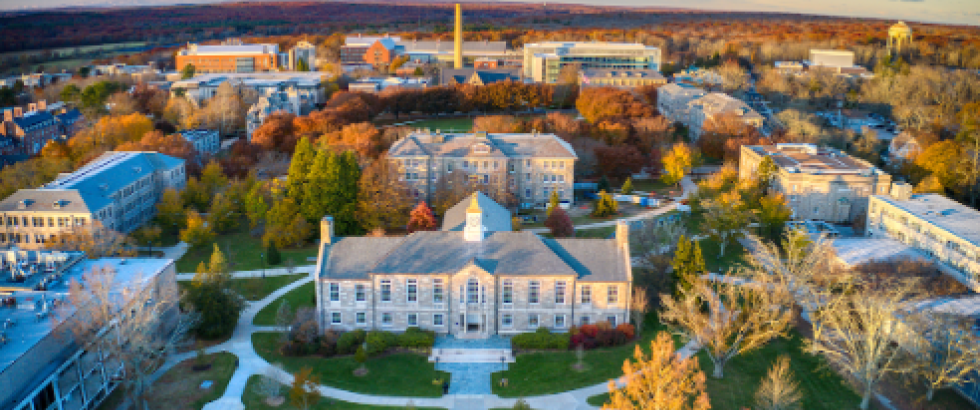 Aerial view of a college campus with multiple academic buildings arranged around lawns and intersecting walkways. Trees across the campus are in autumn foliage, and a large central building with a cupola sits prominently in the foreground.