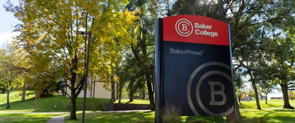 Campus sign reading “Baker College” and “BakerProud” with a large B logo stands on a landscaped brick base beside a sidewalk. Trees with green and yellow leaves surround the sign, and sunlight filters through branches across a grassy campus lawn.