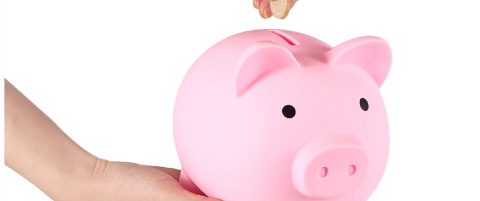 A hand places a coin into the slot of a piggy bank while another hand supports it from below. The piggy bank is smooth and rounded with a simple face design, shown against a plain background.