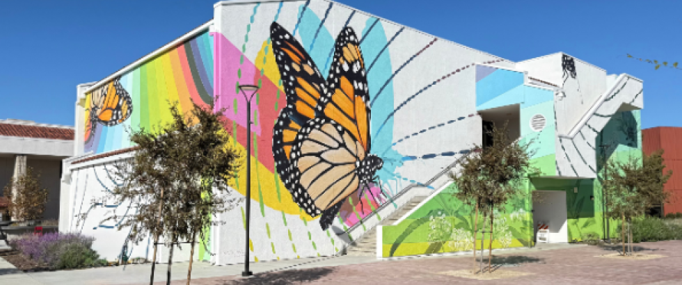A large outdoor mural on a campus building shows several monarch butterflies over colorful rainbow shapes and abstract patterns. The mural wraps around a white building with an exterior staircase, trees, and a brick walkway in front under a clear blue sky.
