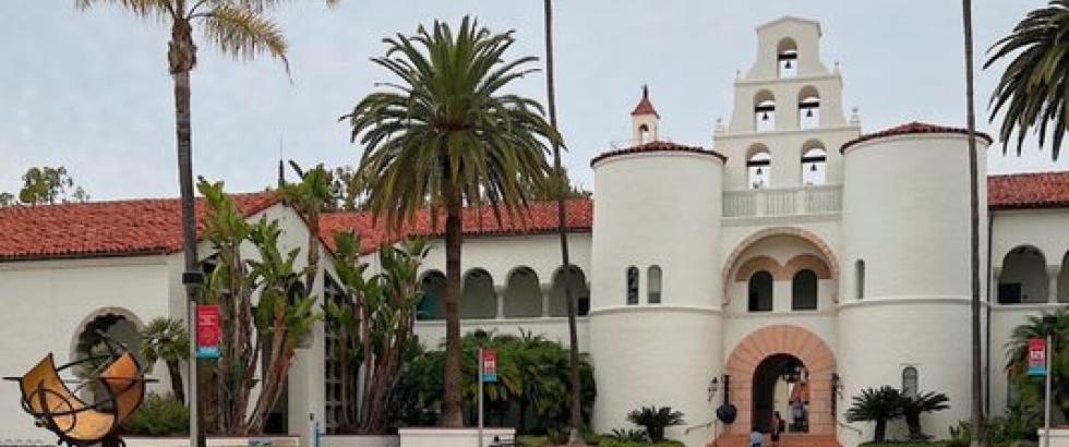 A Spanish-style campus building with white stucco walls, red tile roofs, and a bell tower stands behind a courtyard with palm trees, red flowering shrubs, and a circular brick area featuring a metal sculpture in the foreground.