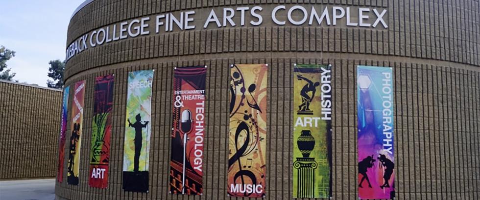 Curved brick building with large metal letters reading “Saddleback College Fine Arts Complex.” Seven vertical banners on the wall read, from left to right: “Dance,” “Art,” “Speech,” “Entertainment & Theatre Technology,” “Music,” “Art History,” and “Photography.” A walkway and small plants are in front of the building.