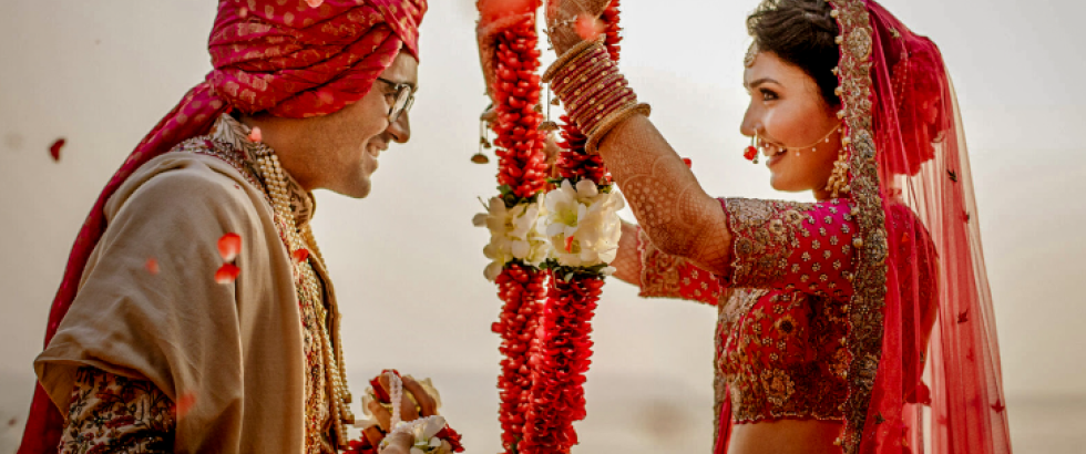 A bride and groom face each other during a wedding ceremony. The bride, wearing a red embroidered outfit with jewelry and a veil, lifts a flower garland to place around the groom’s neck. The groom, wearing a patterned outfit and a red turban, smiles and holds a matching garland. Flower petals fall around them.
