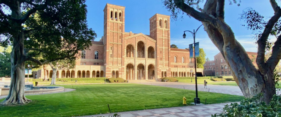 A large brick campus building with two tall towers and a central arched entrance stands beyond a wide green lawn. Trees frame the scene, with a paved walkway, a small fountain to the left, and a person walking near the building under a clear sky.