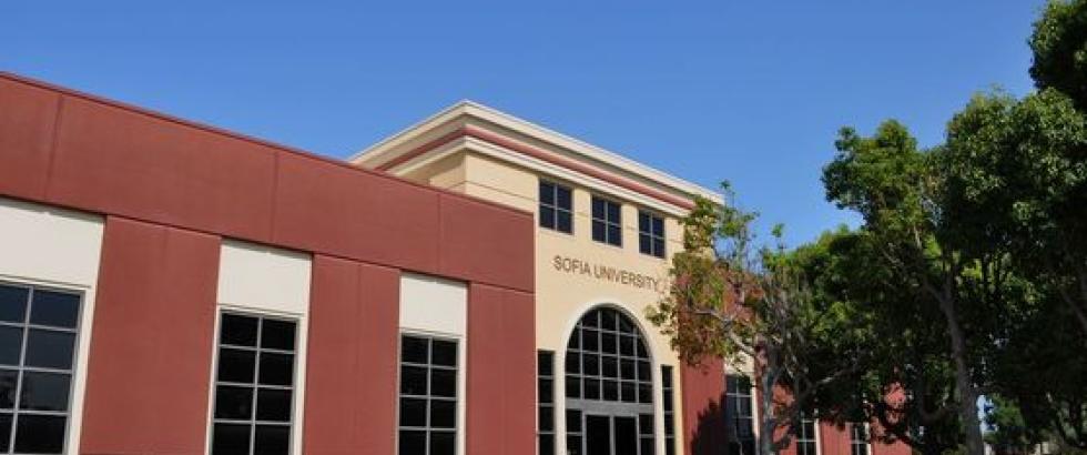 A campus building with a tall arched entrance labeled “Sofia University.” Large rectangular windows line the front, with trees and landscaping along the walkway under a clear sky.