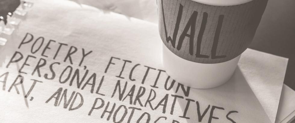 A stack of papers and a coffee cup sit on a table. The cup sleeve reads “WALL,” and the cup is labeled “Literary Journal.” A handwritten sign on paper reads: “Poetry, Fiction, Personal Narratives, Art, and Photography.”