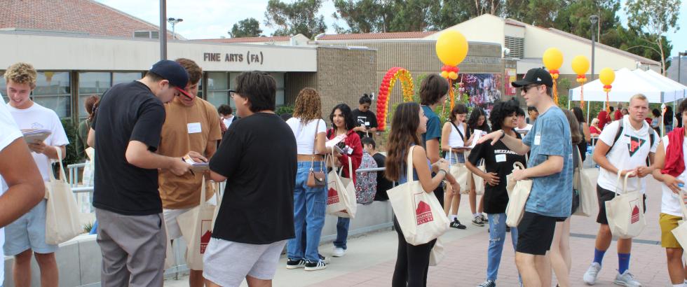 A lively outdoor student event on a college campus plaza, with diverse groups of young adults talking, holding papers, and carrying matching Saddleback College tote bags in front of a "Fine Arts (FA)" building.