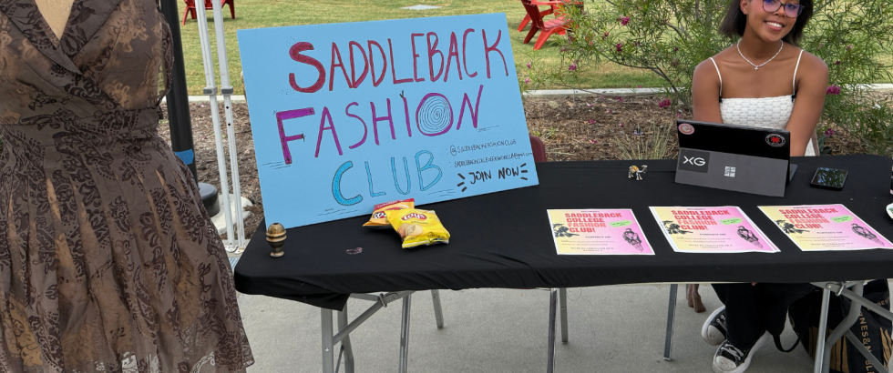 A student sits at a table for the Saddleback Fashion Club. Near her is a a body form with a brown lace dress.