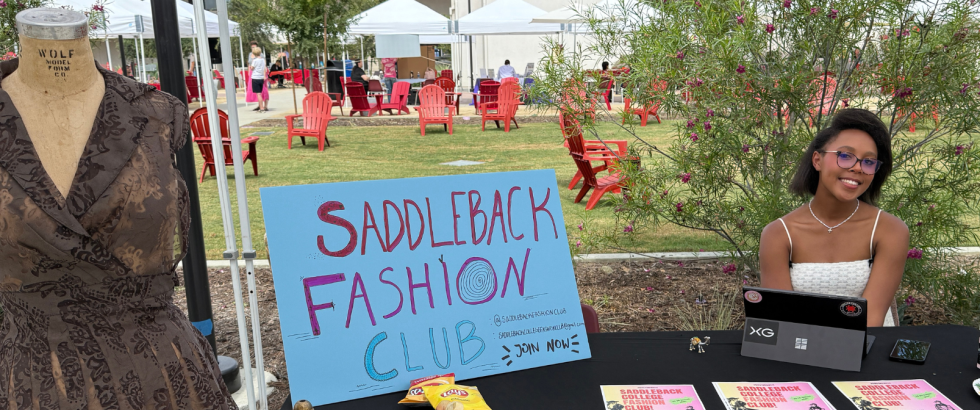 A student sits at a table for the Saddleback Fashion Club. Near her is a a body form with a brown lace dress.