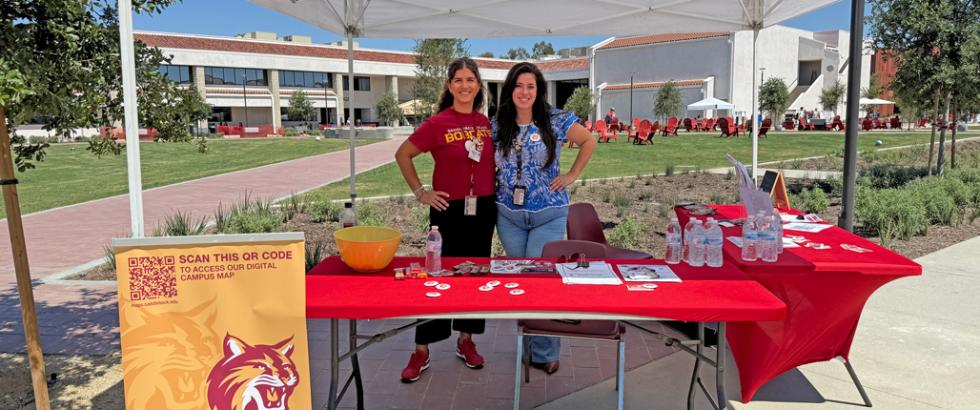 Two female employees of Saddleback College stand at a welcome booth ready to provide information and answer questions.