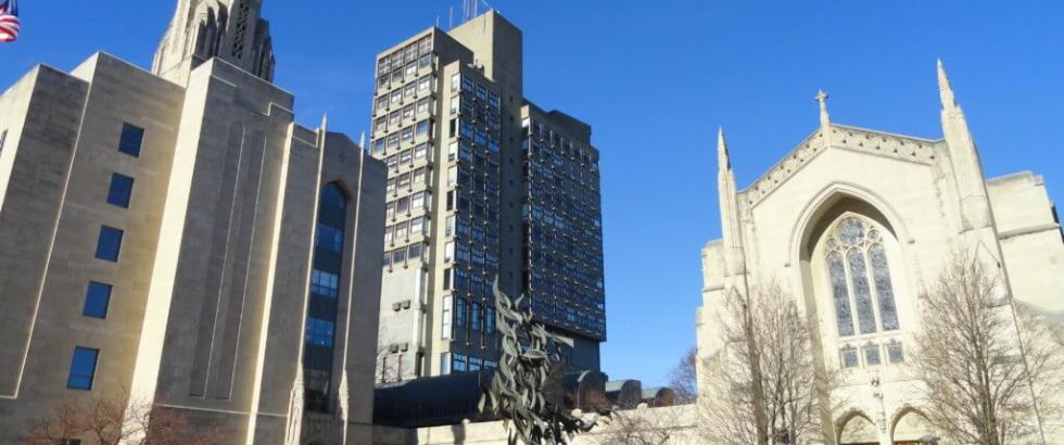 A sunny view across Boston University's Marsh Plaza, featuring the Gothic-style Marsh Chapel, a central bronze sculpture, and campus high-rises under a clear blue sky.