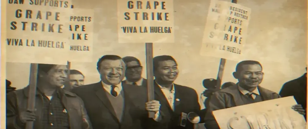 Black-and-white historical photograph of smiling men marching in support of the Delano grape strike. Protesters hold signs reading “UAW Supports Grape Strike,” “UAW President Walter P. Reuther Supports Grape Strike,” and “Viva La Huelga.”