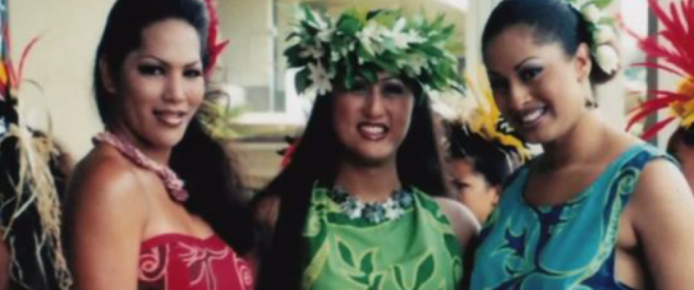 Three smiling women pose together wearing colorful Polynesian-inspired dresses in red, green, and turquoise. The woman in the center wears a leafy floral crown, while the others have flowers in their hair. Hina Wong-Kalu