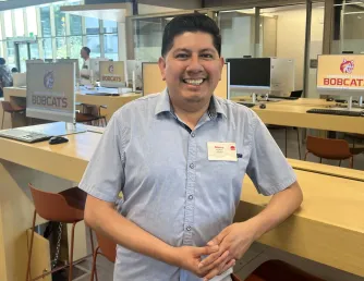 A male Enrollment Support Coach smiling and leaning against a table in front of computer screens in the Saddleback College Gateway building
