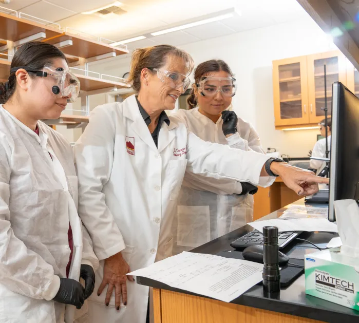 Two female students with female instructor in lab coats and protective goggles reviewing information on a computer screen.